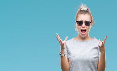 Young blonde woman wearing sunglasses over isolated background crazy and mad shouting and yelling with aggressive expression and arms raised. Frustration concept.