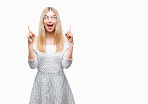 Young beautiful blonde woman wearing glasses over isolated background amazed and surprised looking up and pointing with fingers and raised arms.