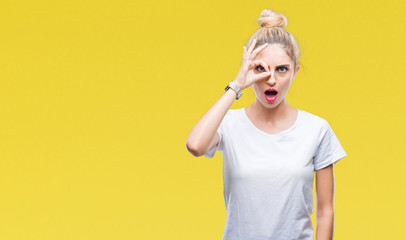 Young beautiful blonde woman wearing white t-shirt over isolated background doing ok gesture shocked with surprised face, eye looking through fingers. Unbelieving expression.