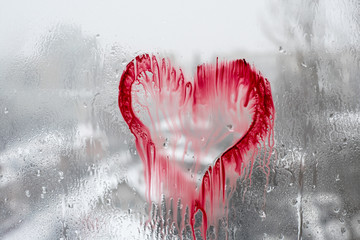 Drops of water on the window pane wash off the painted image of the heart against the background of the snow-covered city