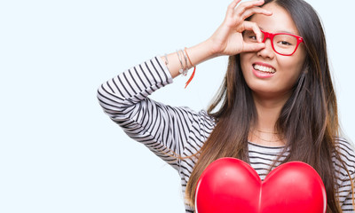 Young asian woman in love holding read heart over isolated background with happy face smiling doing ok sign with hand on eye looking through fingers