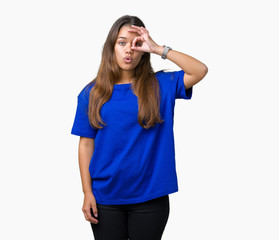 Fototapeta premium Young beautiful brunette woman wearing blue t-shirt over isolated background doing ok gesture shocked with surprised face, eye looking through fingers. Unbelieving expression.
