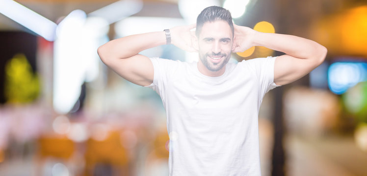 Handsome man wearing white t-shirt over night outdoors background Relaxing and stretching with arms and hands behind head and neck, smiling happy