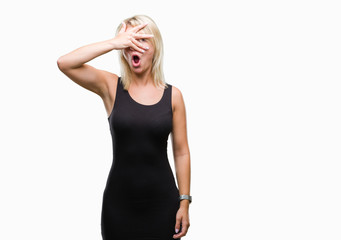 Young beautiful blonde attractive woman wearing elegant dress over isolated background peeking in shock covering face and eyes with hand, looking through fingers with embarrassed expression.