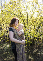 Young redhead babywearing mother wrapping her newborn baby in a sling on a walk in a park on a sunny spring day.
