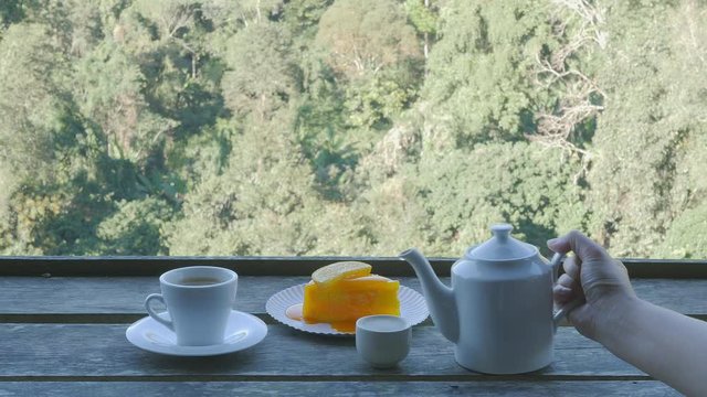 Lady pouring hot tea into a cup in a fresh morning with orange cake and coffee cup over green hill forest background in Chiang Mai Thailand
