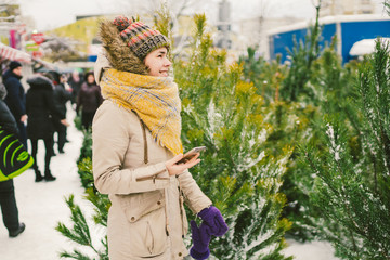 Theme is symbol of Christmas and New Year holidays. Beautiful young Caucasian woman with phone. Hand buyer chooses, makes purchase on Christmas tree market in winter outside in snowy weather