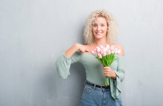 Young blonde woman over grunge grey background holding flowers with surprise face pointing finger to himself