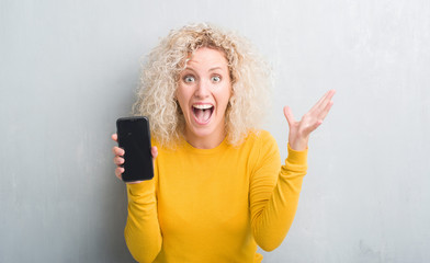 Young blonde woman over grunge grey background showing blank screen of smartphone very happy and excited, winner expression celebrating victory screaming with big smile and raised hands