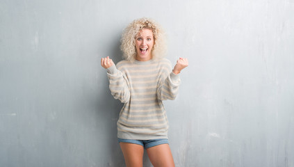 Young blonde woman with curly hair over grunge grey background celebrating surprised and amazed for success with arms raised and open eyes. Winner concept.