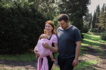 Obraz premium Young parents on a walk with their baby wrapped in a pink sling.