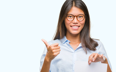 Young asian woman over isolated background holding blank paper happy with big smile doing ok sign, thumb up with fingers, excellent sign