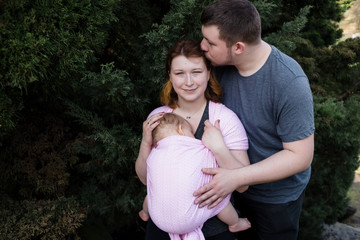 Young parents with baby in a sling outside. Husband kissing his wife on her forehead.