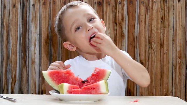 The child sits at the table on the background of a wooden wall and with pleasure eats a watermelon.