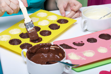 A woman using a brush brushes a silicone dessert mold with a melted black chocolate. Nearby are other ingredients.