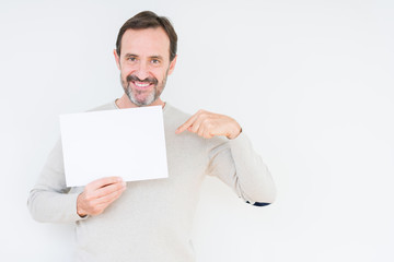 Senior man holding blank paper sheet over isolated background with surprise face pointing finger to himself