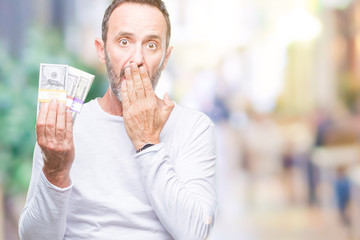 Senior hoary man holding buch of dollars over isolated background cover mouth with hand shocked with shame for mistake, expression of fear, scared in silence, secret concept