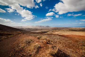 Amazing vulcanic landscape with mountains, ocean and small village Lajares on horizon. Sky is blue with clouds, gravel is red and brown. Fuerteventura Island, Canary Island, Spain, Europe.
