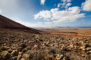 Amazing vulcanic landscape with mountains, ocean and small village Lajares on horizon. Sky is blue with clouds, gravel is red and brown. Fuerteventura Island, Canary Island, Spain, Europe.