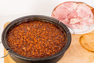 Southern Style Baked Beans in Dutch Oven in Front of Baked Ham on White BG.