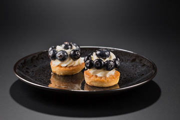 Two small cakes with blueberries and creamy cream on a black plate.