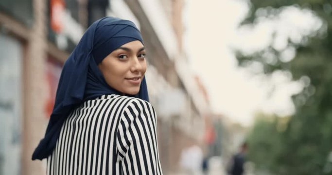 Attractive muslim girl wearing hijab walking down the street, talking with someone on phone, twirling around and looking at camera - modern muslim, modern lifestyle concept closeup 4k