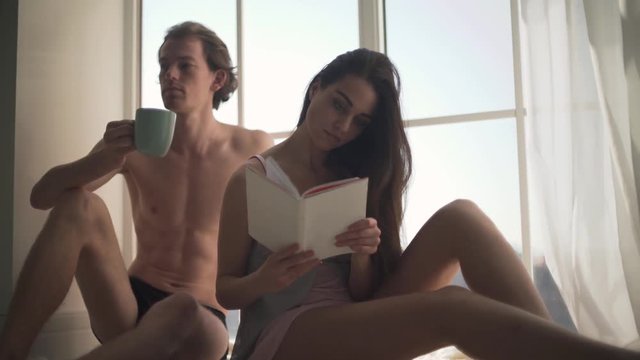 Young half-naked couple spends leisure in the morning. Beautiful sexy woman in nightie reading a book with young naked man who drink cup of coffee near window from floor-to-ceiling.