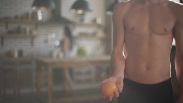 Guy with a naked torso throws an orange while standing in the kitchen. Girl in the background is cooking.