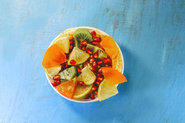 cut scitrus fruits in a white plate. orange, pomegranate, mandarin, persimmon, kiwi. on a blue wooden board.