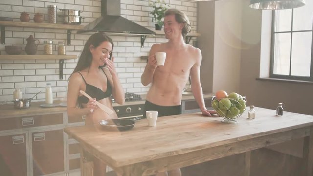 Smilling young naked couple in the beautiful loft kitchen. Joyful guy drinking morning coffee in underwear and a happy girl in lingerie cooking while standing near the kitchen table in the sunlight.