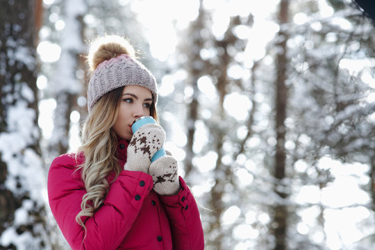 Pretty woman in burgundy coat hold blue thermo cup and drink hot coffee or tea and warm up in winter forest. Winter holiday concept - Image
