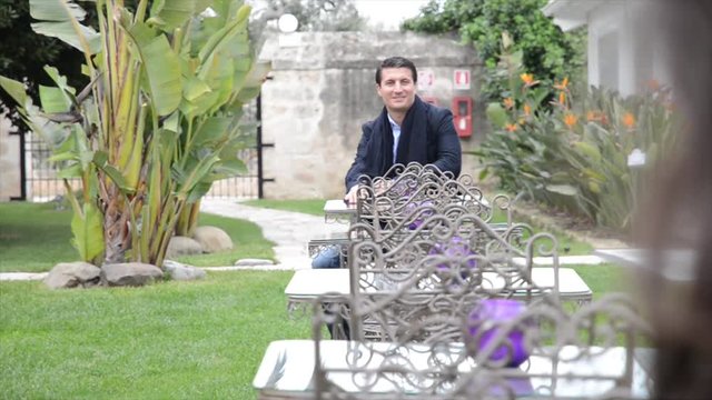 Giovane uomo si siede ad un tavolino in un giardino, osservato da una donna interessata