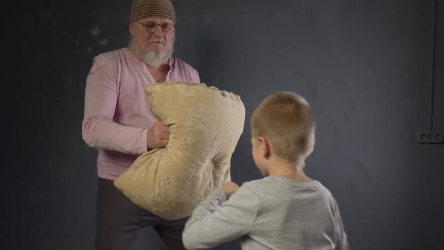 Concept of gymnastics and sport at home. Elderly retired father with gray beard and in pink T-shirt holds pillow and trains boy for boxing. Brave child beats on pillow with all his might.