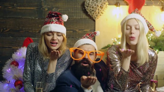 Bearded man in big glasses and two prety girls in New Year's hats blow candy from their palms on the background of the new year tree. Concept of new year mood. New Year gifts.