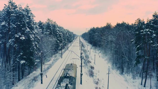 Aerial following shot of the commuter train through the winter snowy forest at sunset