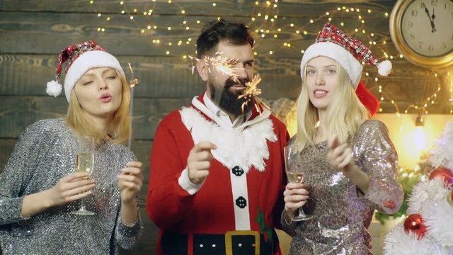 Bearded man in santas costume and two prety girls in New Year's hats burn sparkler lights and drink champagne on the background of the new year. New Year party. New Year mood concept.
