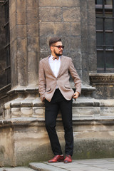 Obraz premium A young bearded boy with a modern hairstyle in a suit and bow tie poses on the camera. Man keeps hands in pockets and looking forward