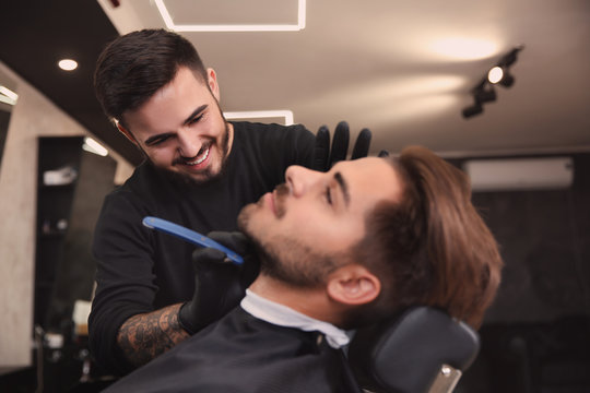 Professional hairdresser shaving client with straight razor in barbershop