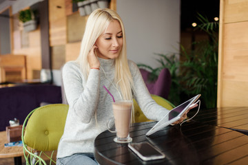 Young blond Woman using Tablet and Drinking Coffee. Relaxing in Cozy cafe, Sit on the Chair and listening with pleasure in headphones an audio book or music