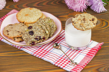 Homemade cookies: coconut, oatmeal, chocolate, on a wooden table, with a glass of milk, against the background of a summer composition with flowers