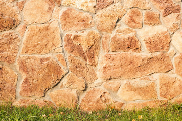 Decorative beige facing stone on the wall and green grass at the bottom of the frame. The texture of the wall of the old stone. Background and wallpaper.