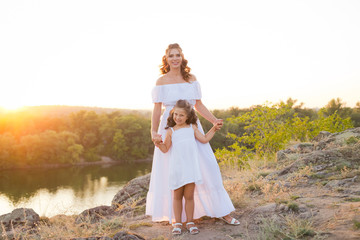 A small smiling laughing girl with curly brown hair dressed white short dress with young beautiful mother in long white dress on the stones at the river on sunset time