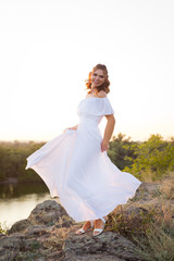 A young smiling laughing woman with curly brown hair dressed in white long dress dancing on the stones at the river on sunset time