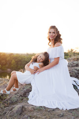 A small smiling laughing girl with curly brown hair dressed white short dress with young beautiful mother in long white dress sitting on the stones at the river on sunset time