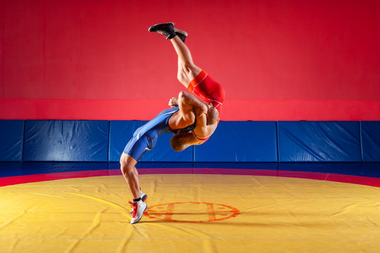 Two strong wrestlers in blue and red wrestling tights are wrestlng and making hip throw wrestling on a yellow wrestling carpet in the gym. Young man doing grapple.