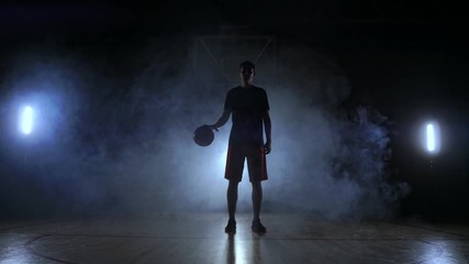 Basketball player in sportswear red shorts and a blue t-shirt goes on a dark basketball court in the backlighting coming out of the smoke knocks a basketball ball on the floor looking at the camera in - Powered by Adobe