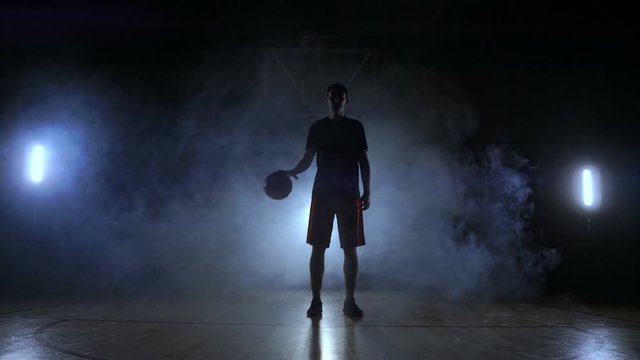 Basketball player in sportswear red shorts and a blue t-shirt goes on a dark basketball court in the backlighting coming out of the smoke knocks a basketball ball on the floor looking at the camera in