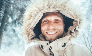 Man dressed in Warm Hooded Casual Parka Jacket Outerwear walking in snowy forest cheerful smiling face portrait. Outdoor time and winter outfit concept image. © Soloviova Liudmyla