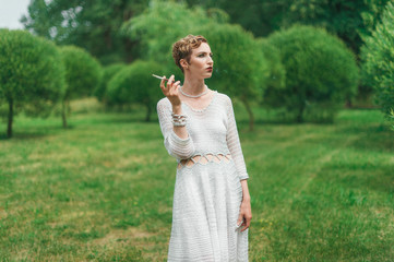 Fototapeta premium Colorful photography of young smoking woman wearing knitted long white dress. Outdoors image