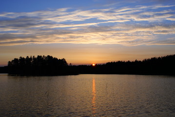 Fototapeta na wymiar Sunrise on the lake. On the horizon a dark forest. The sun appeared over the tops of the trees and painted the sky pink. Blue sky. The water in the lake is dark blue. In the water trail of the sun.